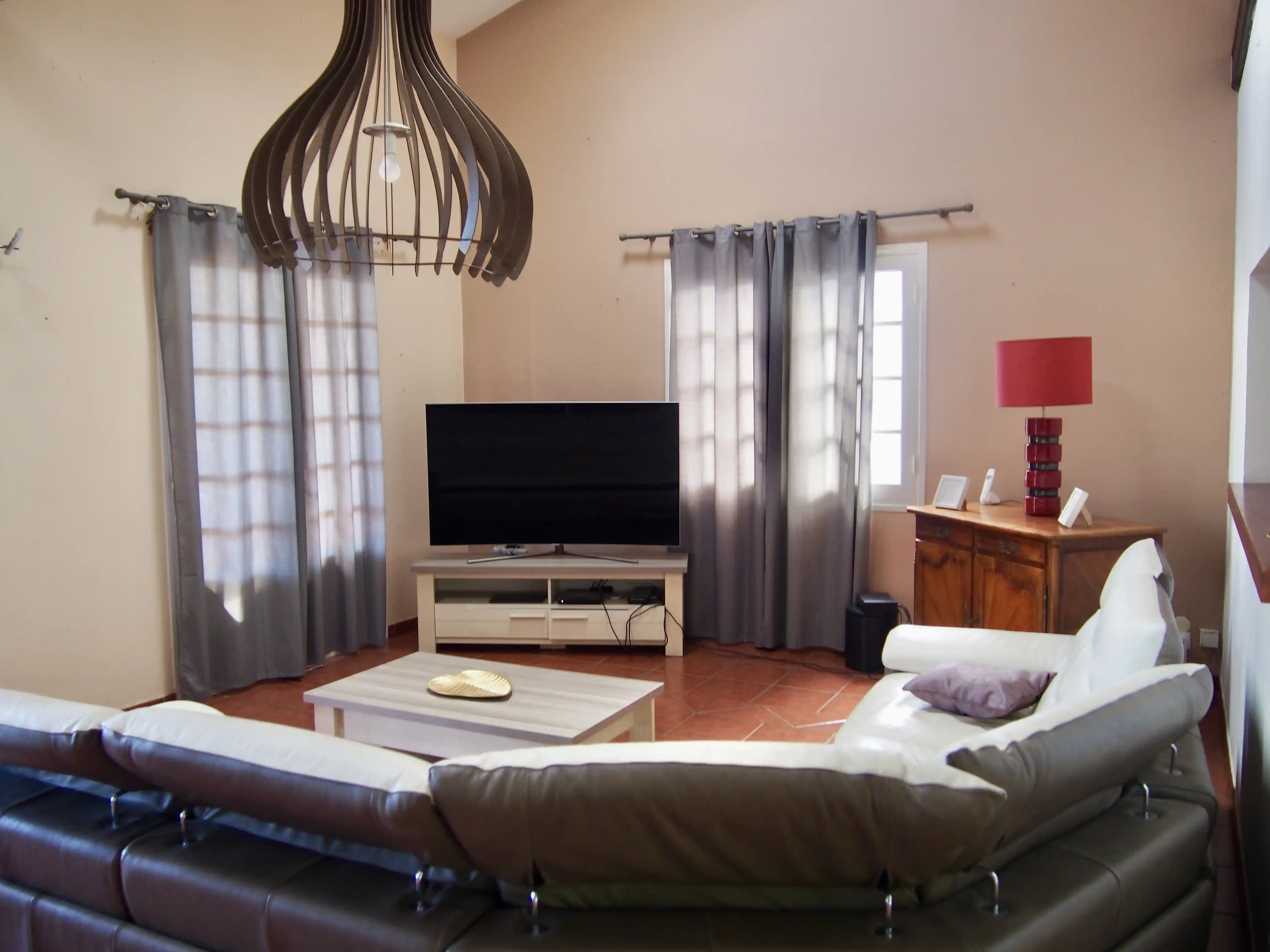 Modern living room with sectional sofa, flat-screen TV on wooden stand, coffee table, and wooden cabinet with red lamp.