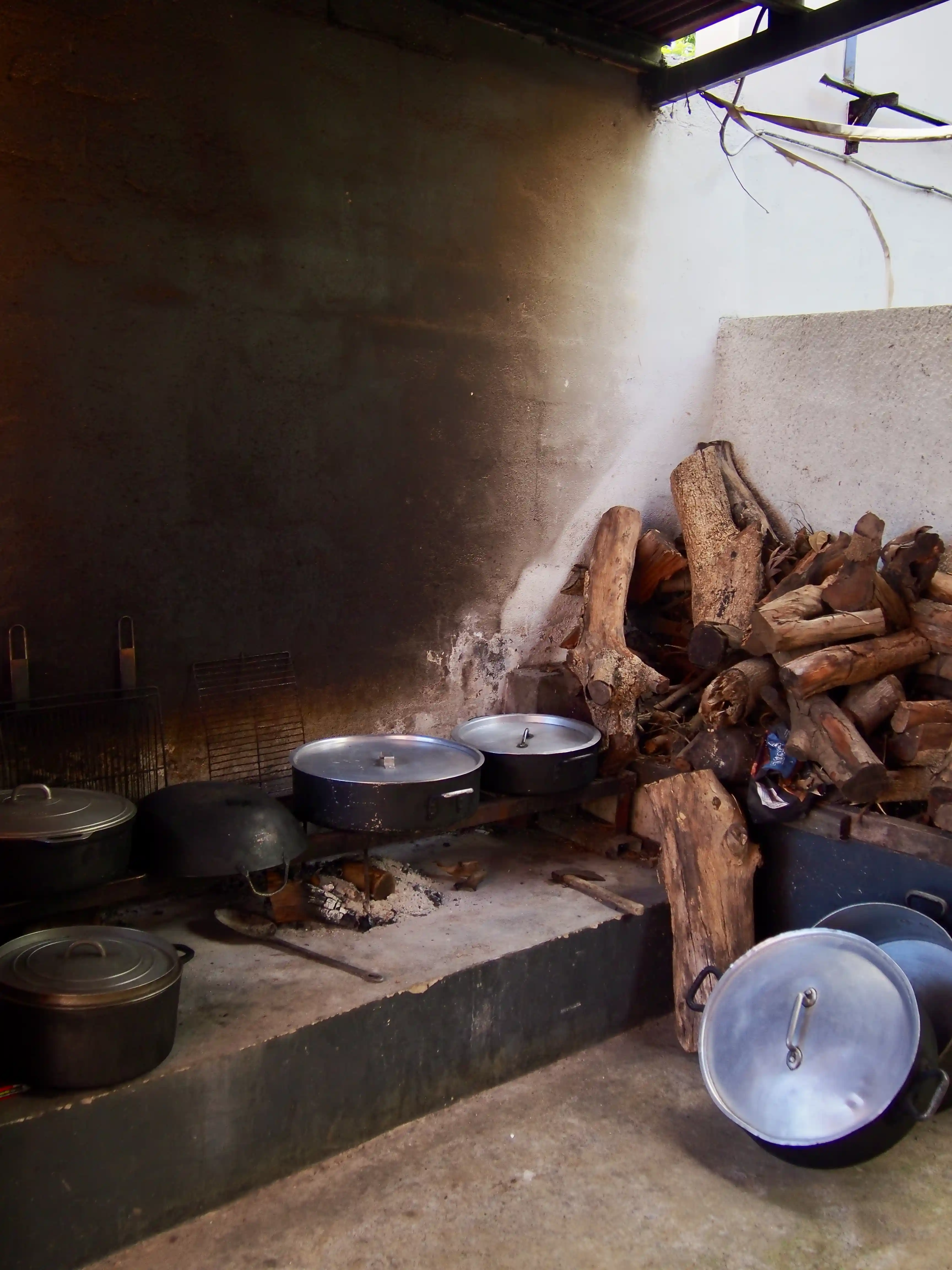 Traditional indoor wood-fired stove with cooking pots on it and a large pile of firewood stacked against the wall.