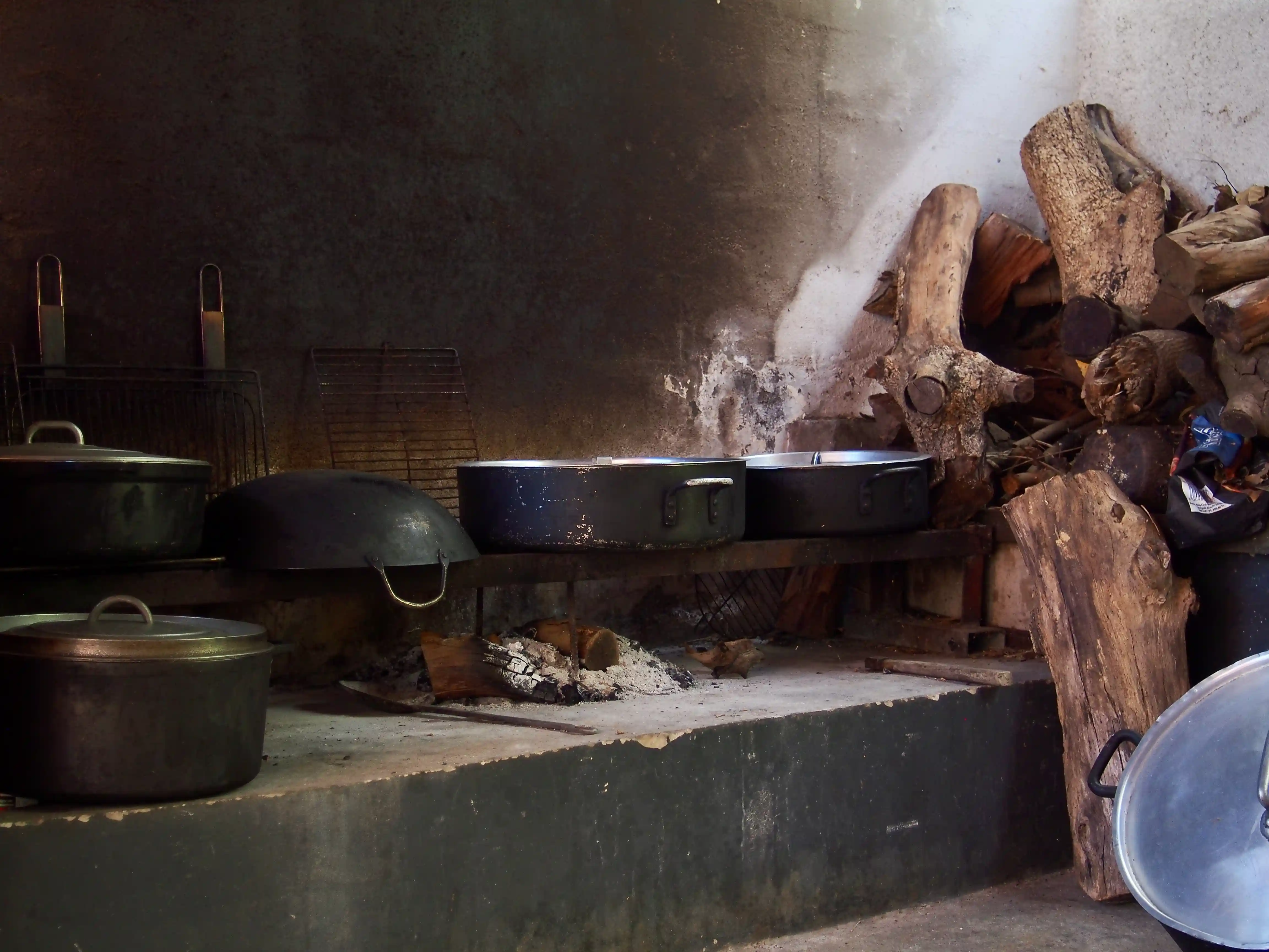 Old wood-burning stove setup with metal pots and pans on a concrete hearth, and a stack of firewood beside it.