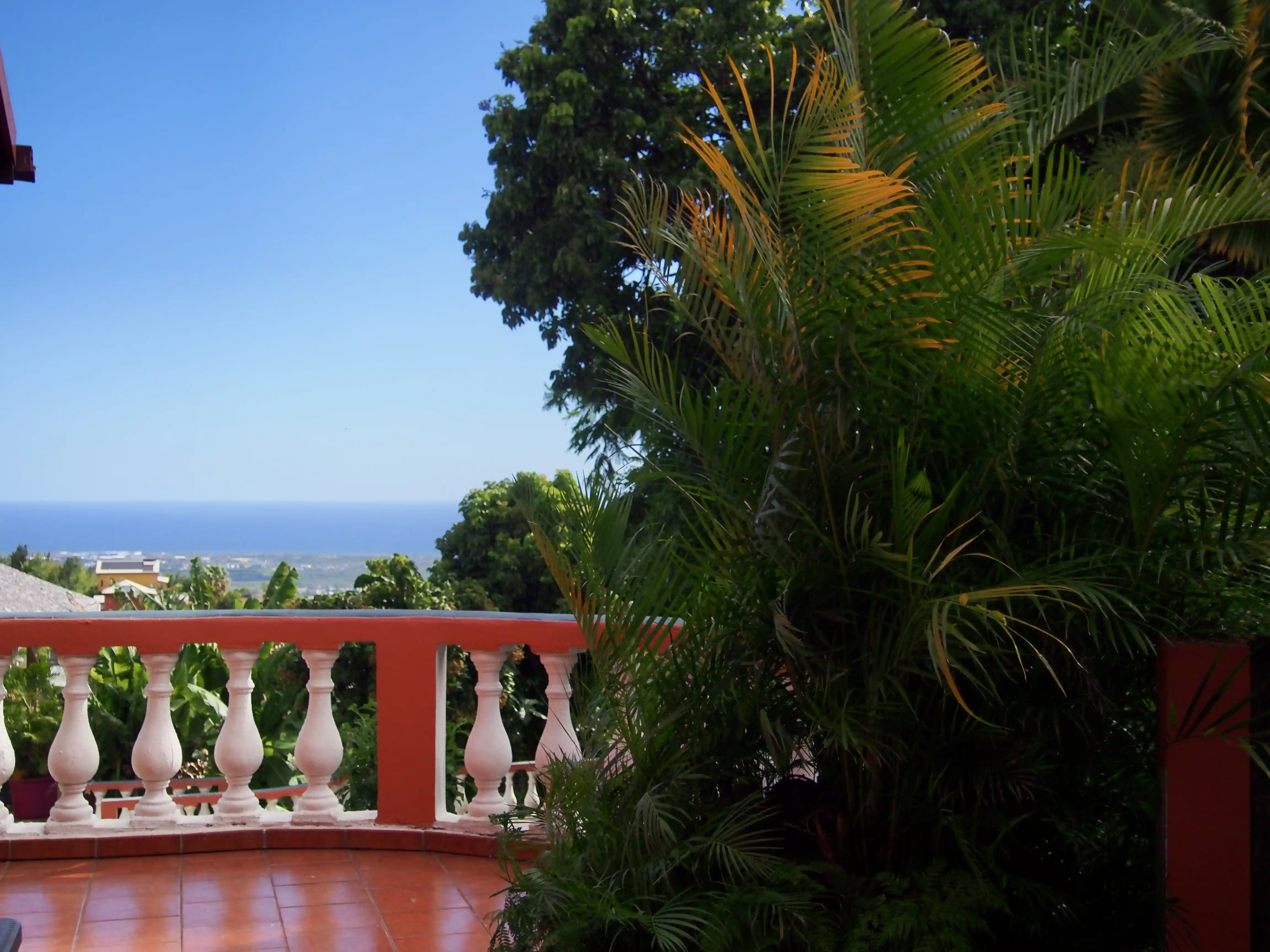 View from a terrace with orange tiles and white balustrade overlooking green trees and a distant ocean under a clear blue sky.