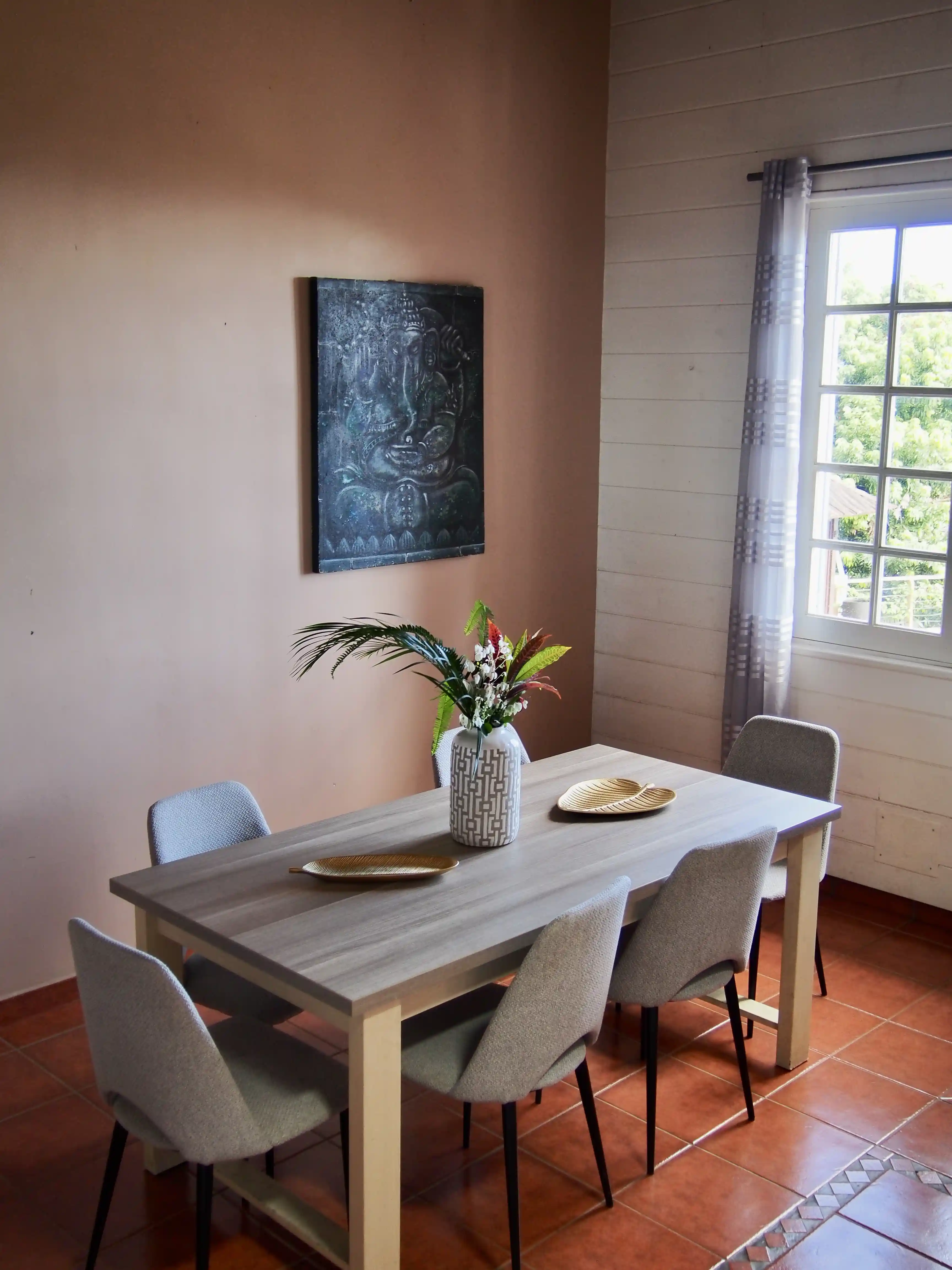 Dining room with a wooden table surrounded by six gray upholstered chairs, a vase with colorful flowers in the center, and a window with a sheer curtain.