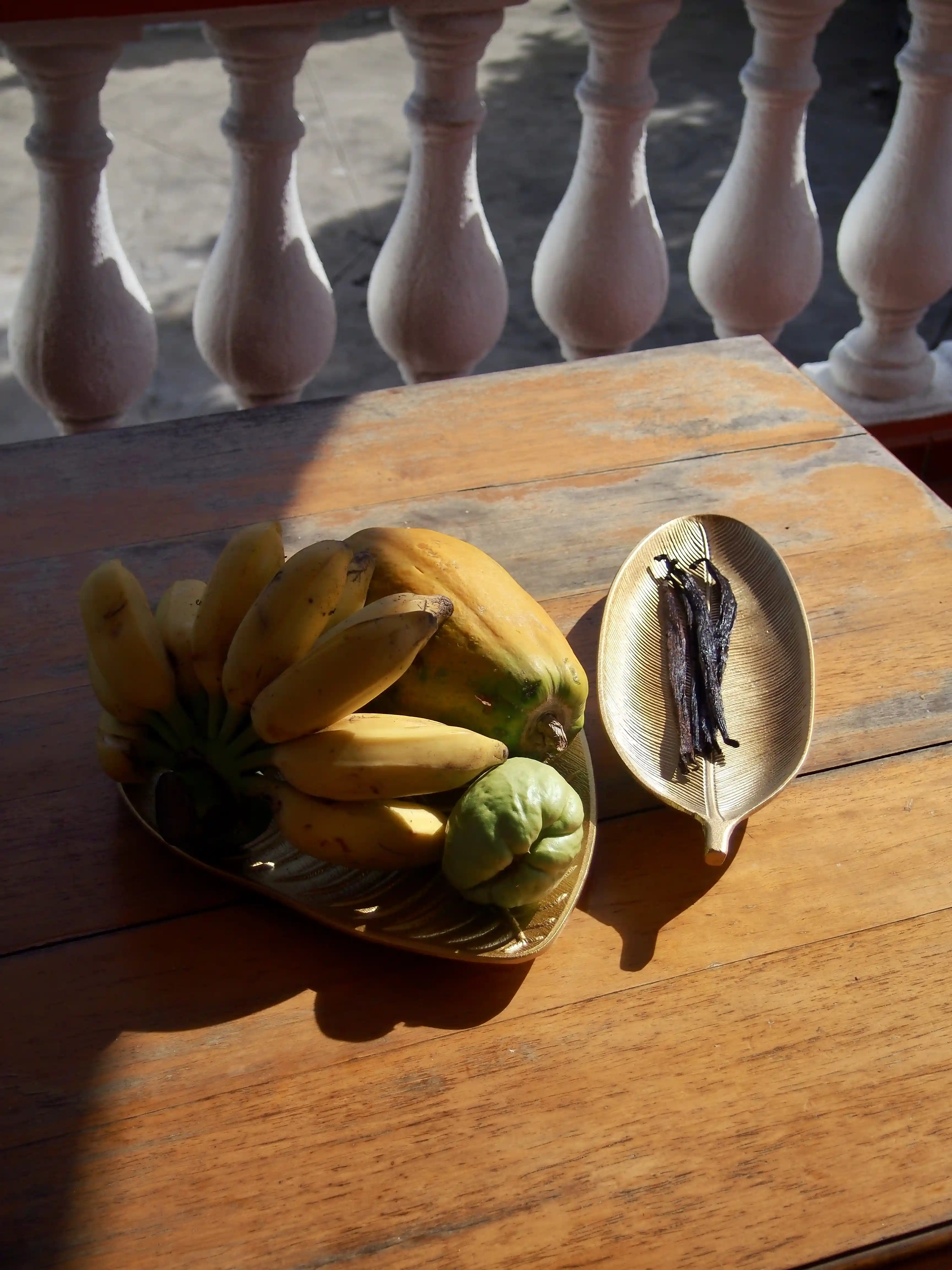 Two decorative plates on a wooden table, one holding bananas, a papaya, and a green fruit, the other holding dried vanilla pods, with stone balusters in the background.
