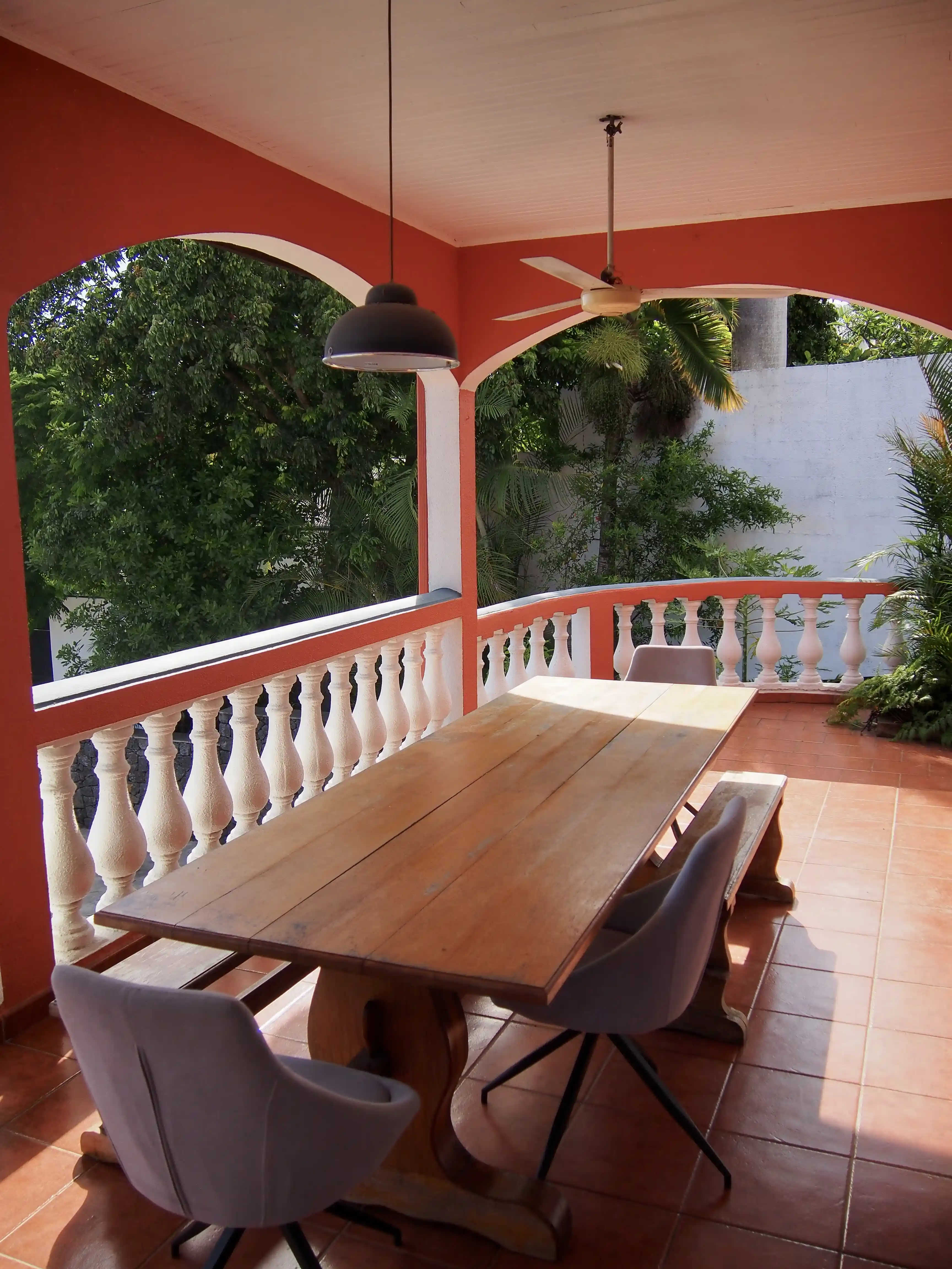 Covered outdoor patio with a wooden dining table, cushioned chairs, bench seating, and a ceiling fan under red arches and surrounded by green foliage.