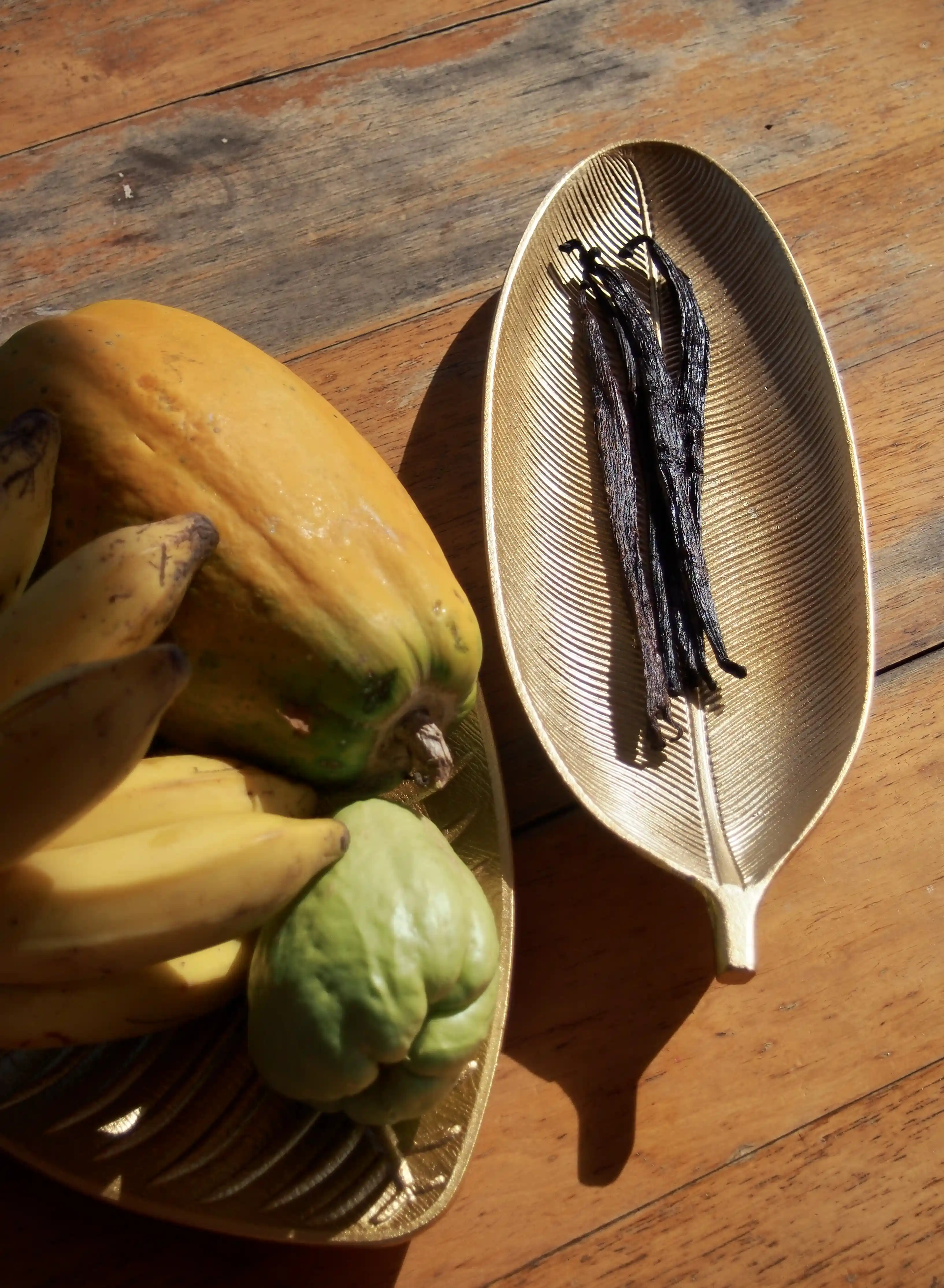 Vanilla beans on a gold leaf-shaped dish next to a gold tray with bananas, papaya, and chayote on a wooden surface.