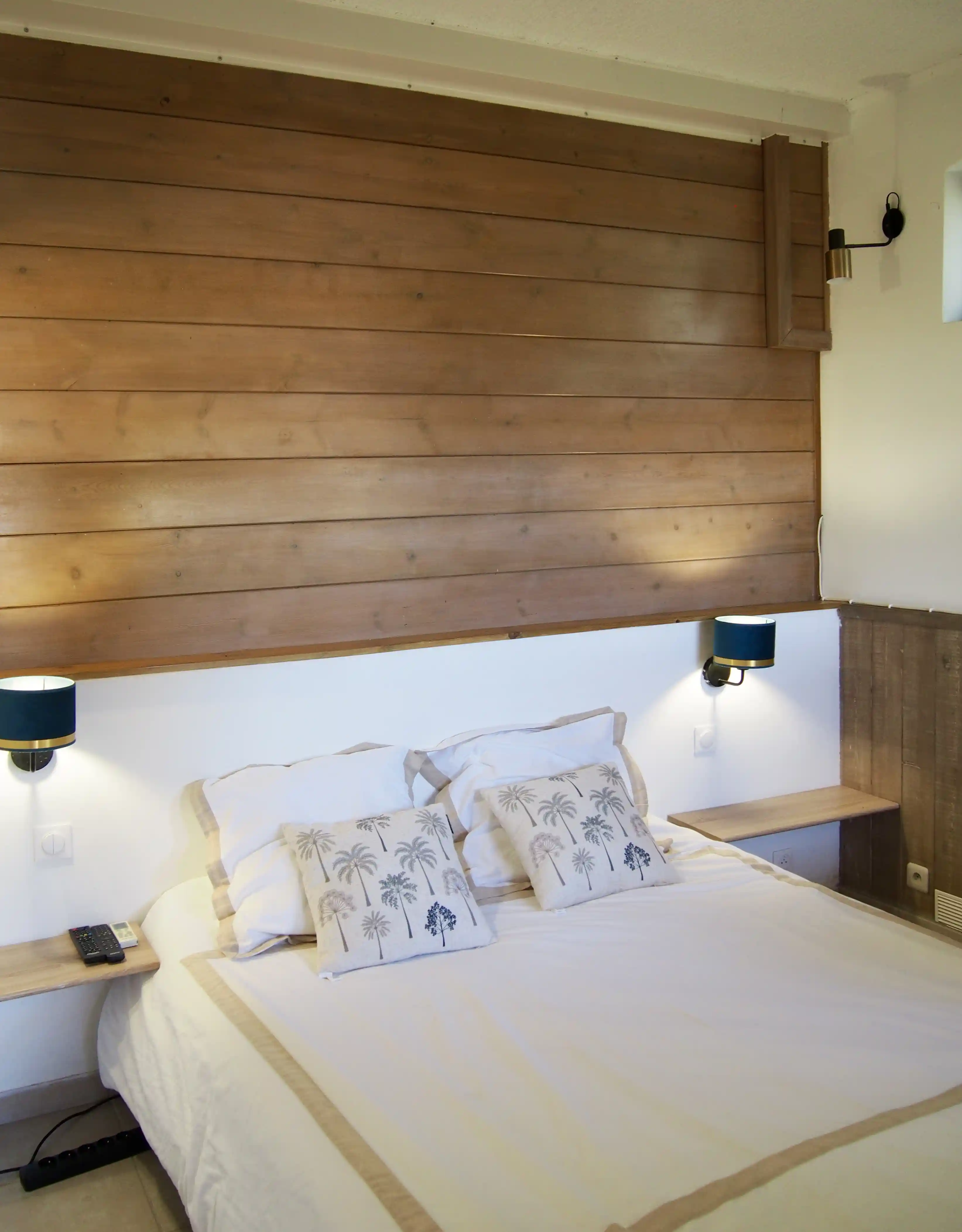 Bed with white and beige bedding and palm tree printed pillows against a wooden paneled wall with blue wall lamps on both sides.