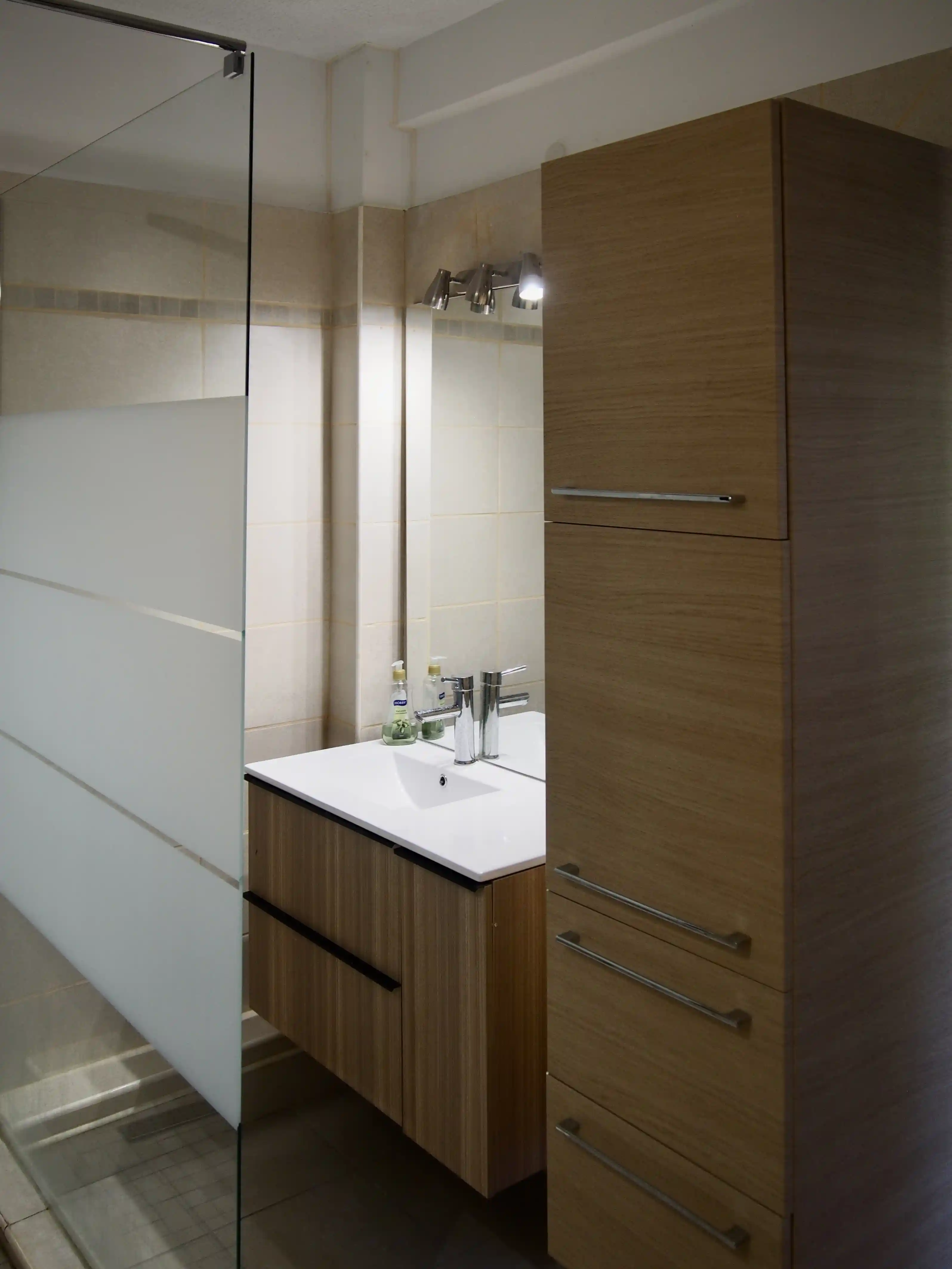 Modern bathroom with wood-finished vanity, white sink, tall wooden cabinet, and frosted glass shower door.