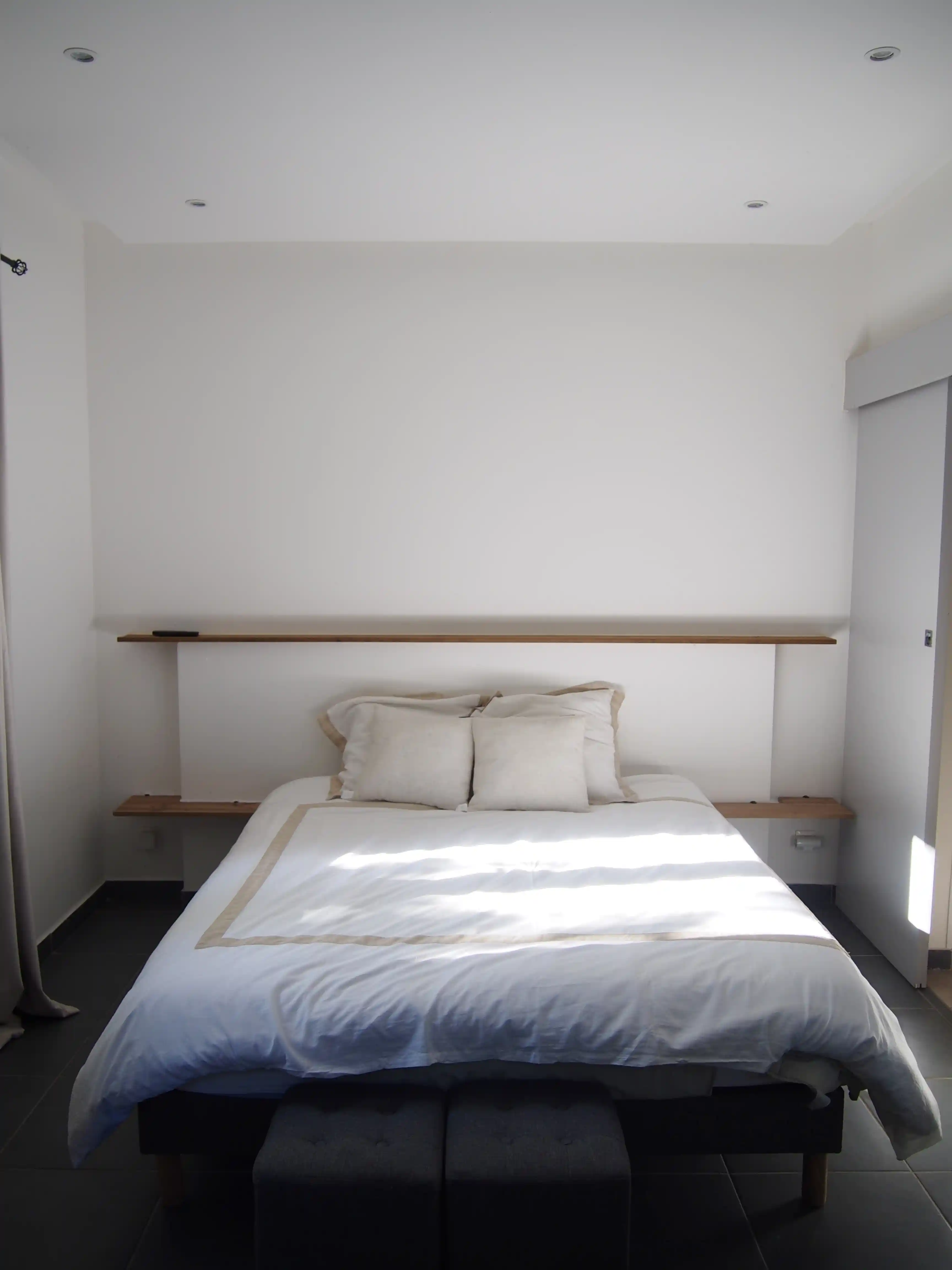 Minimalist bedroom with a white bed, beige pillows, two wooden shelves on the wall, and dark tile flooring.