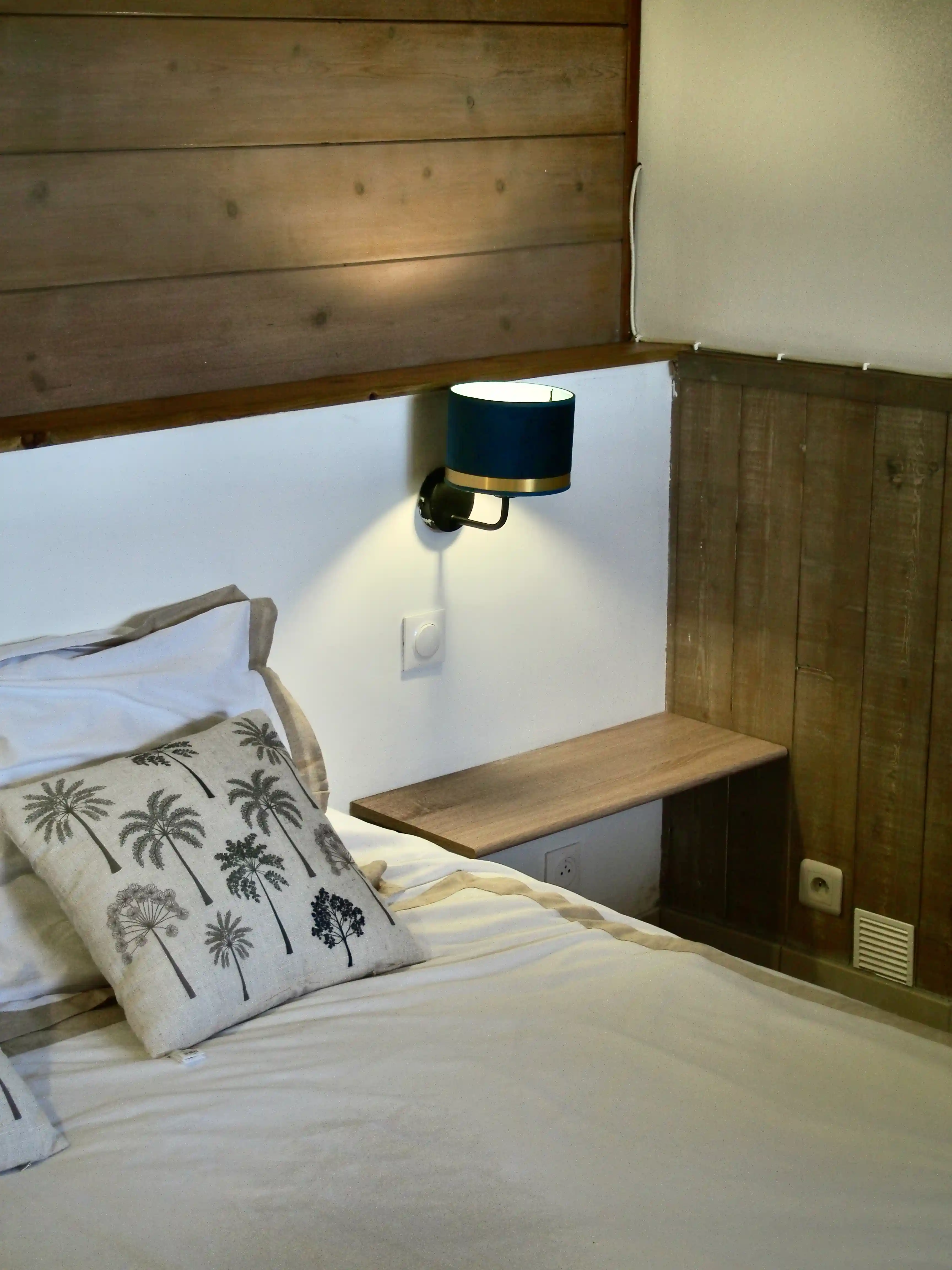 Bed with white bedding, palm tree patterned pillow, wooden walls, and a wall-mounted blue lamp.