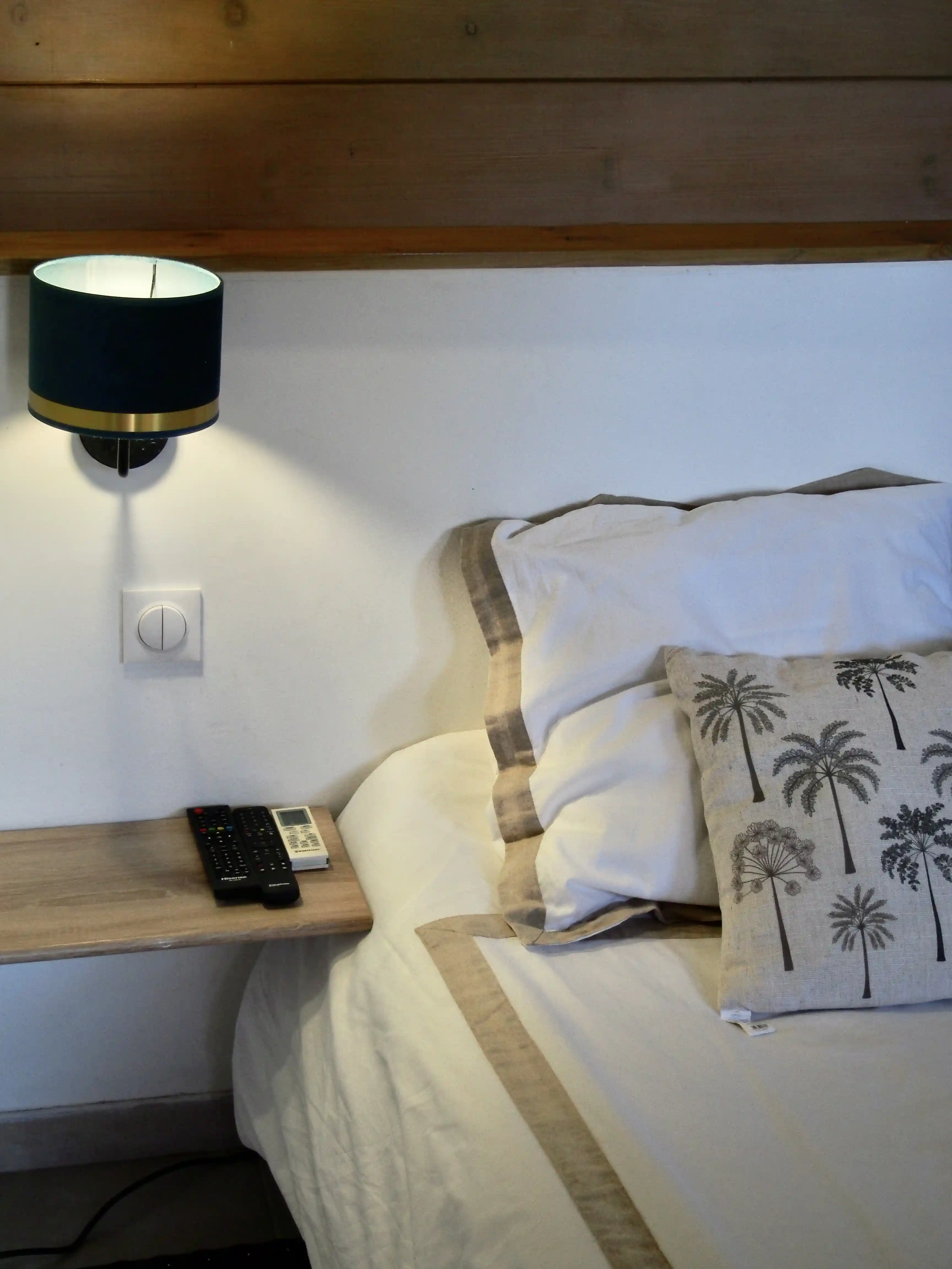Side view of a bed with white linens bordered by beige stripes, a decorative pillow with palm tree prints, a wooden bedside shelf with three remote controls, and a wall-mounted black and gold lamp.