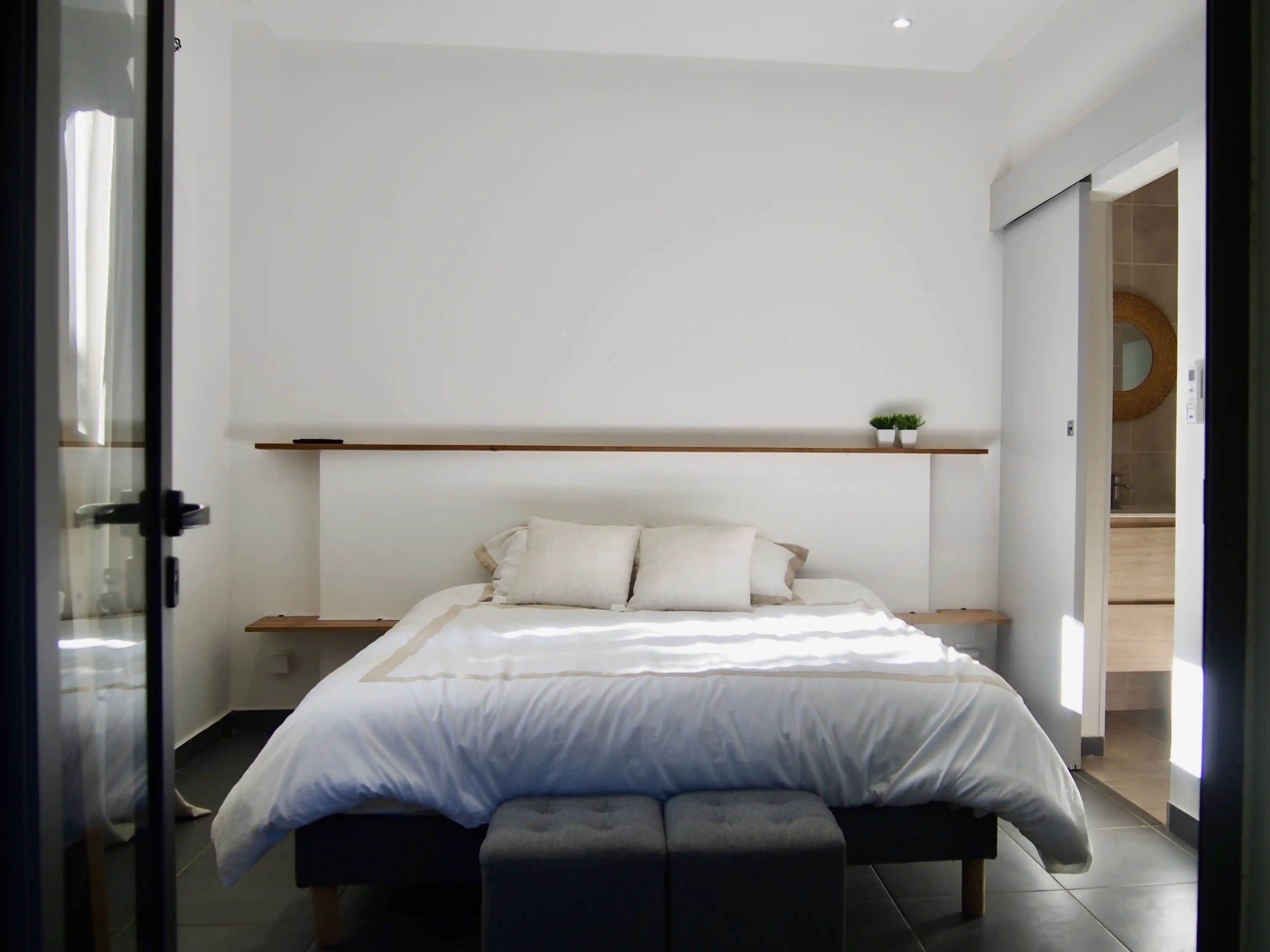 Minimalist bedroom with a double bed, white bedding, two beige pillows, wooden shelves with small plants, and an open door leading to a bathroom.