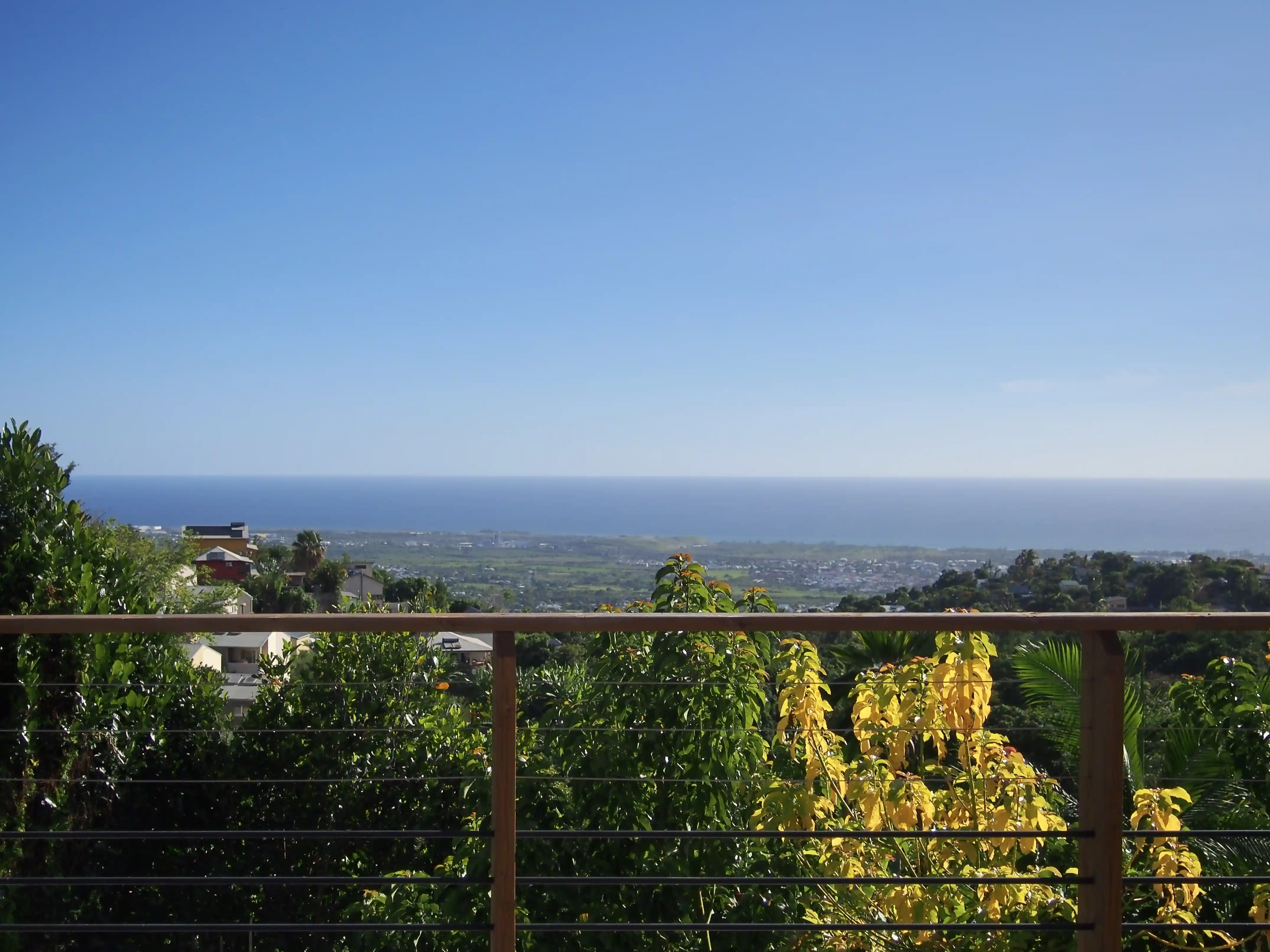 View from a balcony over lush greenery and houses toward the distant ocean under a clear blue sky.