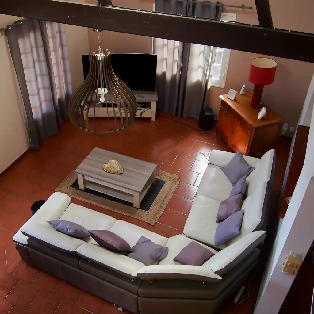 Cozy living room with an L-shaped white sofa, gray and purple pillows, wooden coffee table on a woven rug, a TV, and a red table lamp on a wooden cabinet.