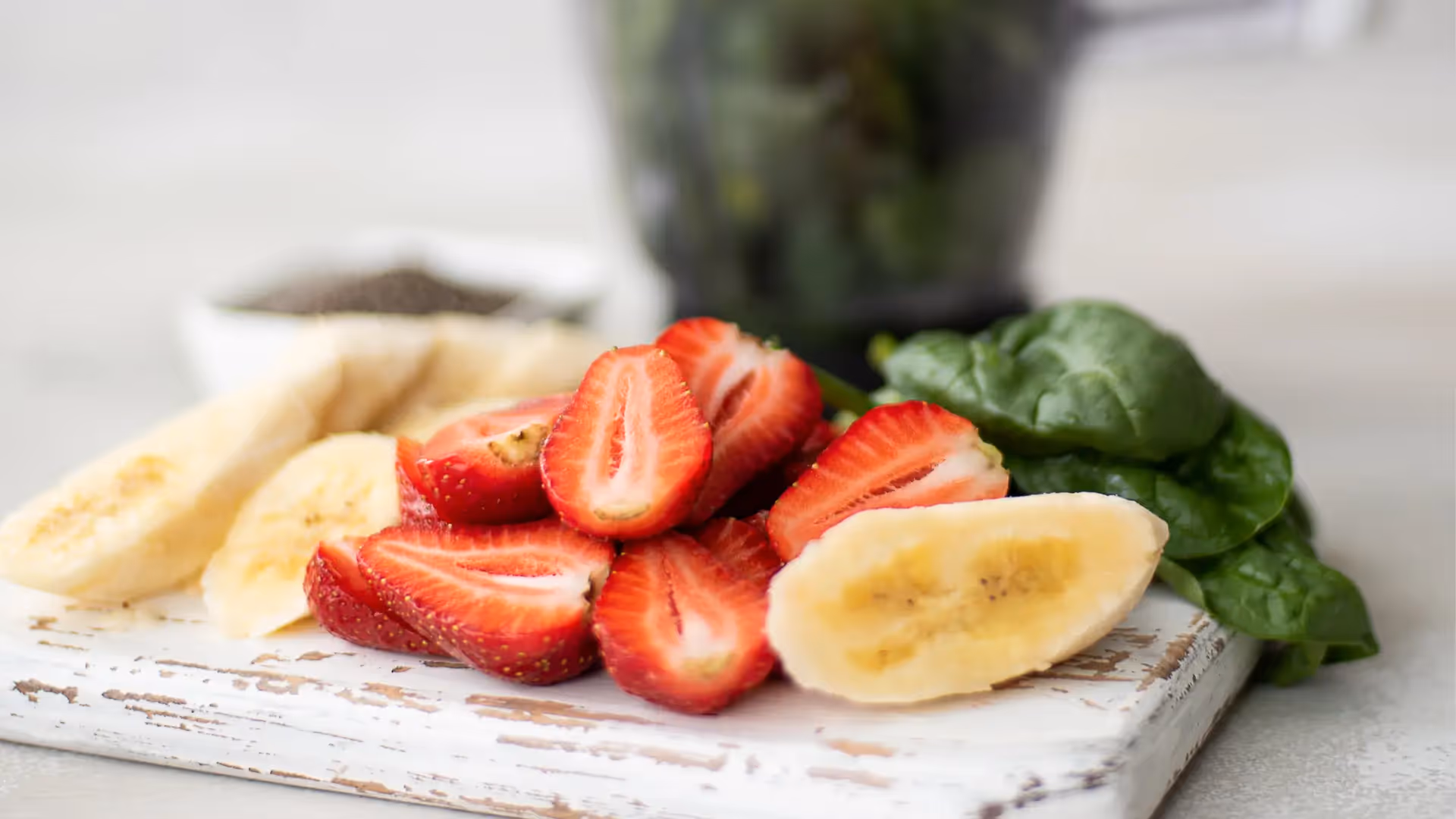 Smoothie ingredients on a counter: a carton of oat milk, a bowl of spinach, a bowl of frozen berries, and sliced bananas on a cutting board, with a window view in the background.
