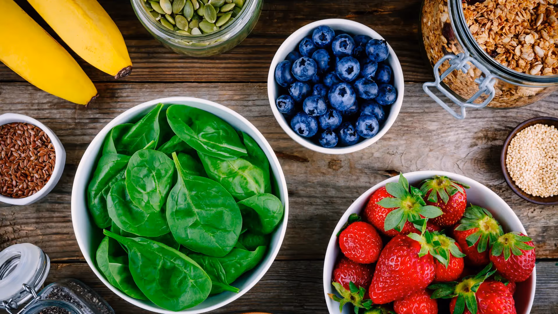 Smoothie ingredients on a counter: a carton of oat milk, a bowl of spinach, a bowl of frozen berries, and sliced bananas on a cutting board, with a window view in the background.