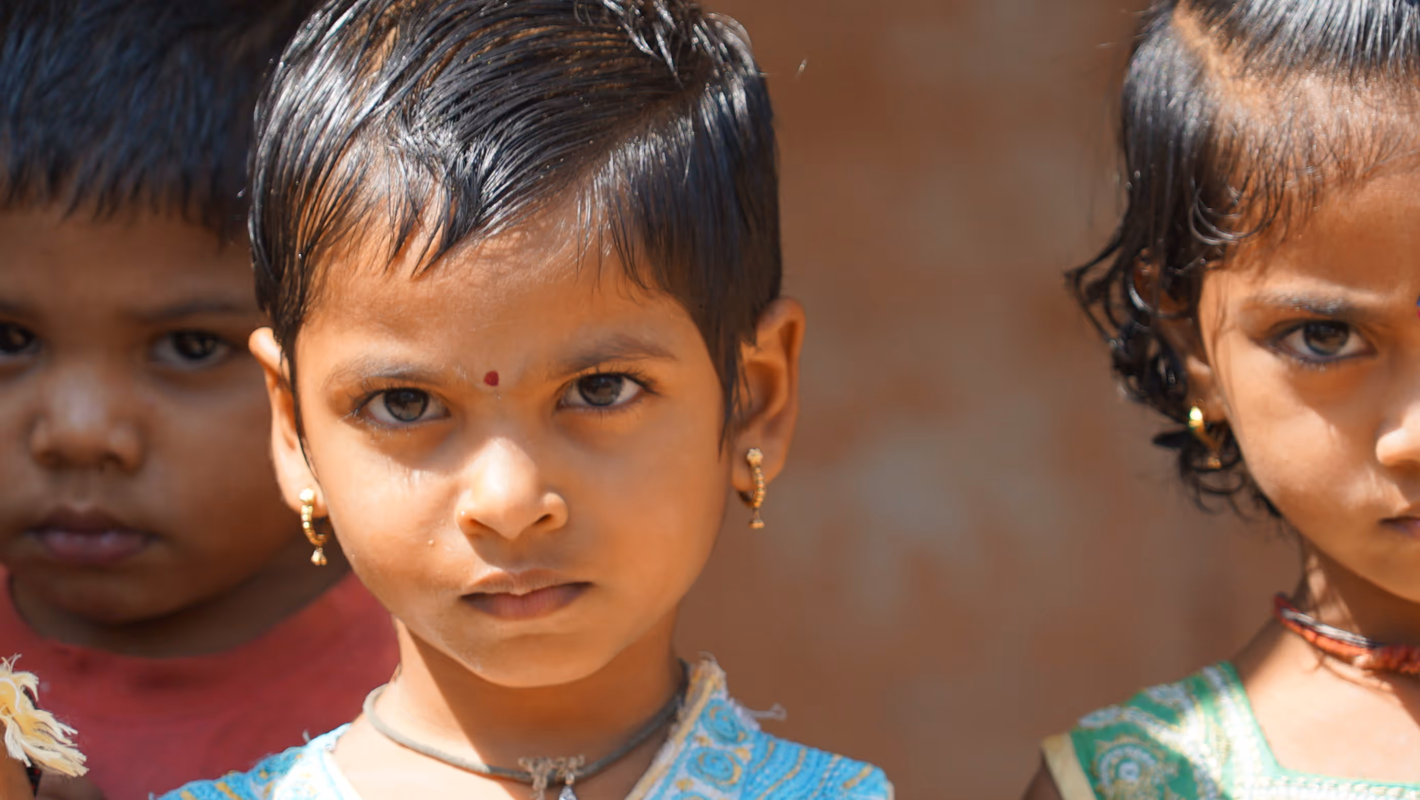 Drei Kinder mit ernstem Gesichtsausdruck im Sonnenlicht, zwei Mädchen tragen traditionelle Ohrringe.
