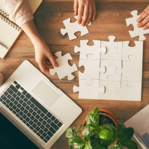 Two people assembling white puzzle pieces on a wooden table with a laptop, notebook, and a green plant nearby.