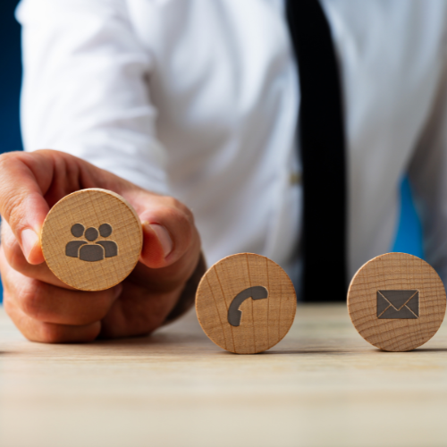 Hand holding a wooden block with a group of people icon, next to blocks with phone and envelope icons on a wooden table.