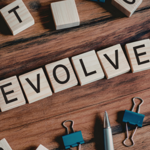 Wooden letter tiles spelling the word EVOLVE on a wooden surface with binder clips and a pen nearby.
