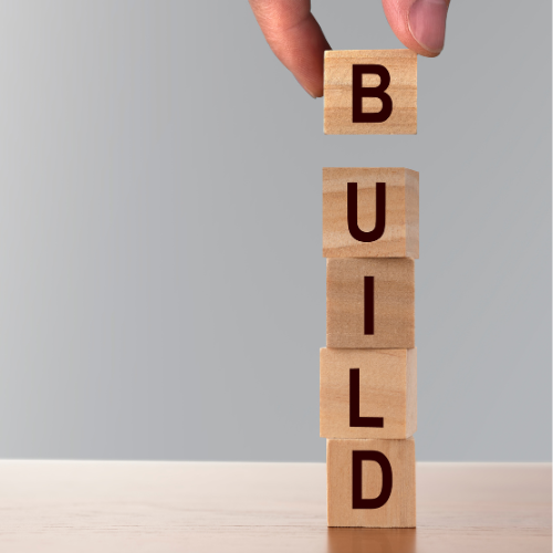 Hand placing a wooden block with the letter B on top of stacked wooden blocks spelling the word BUILD.