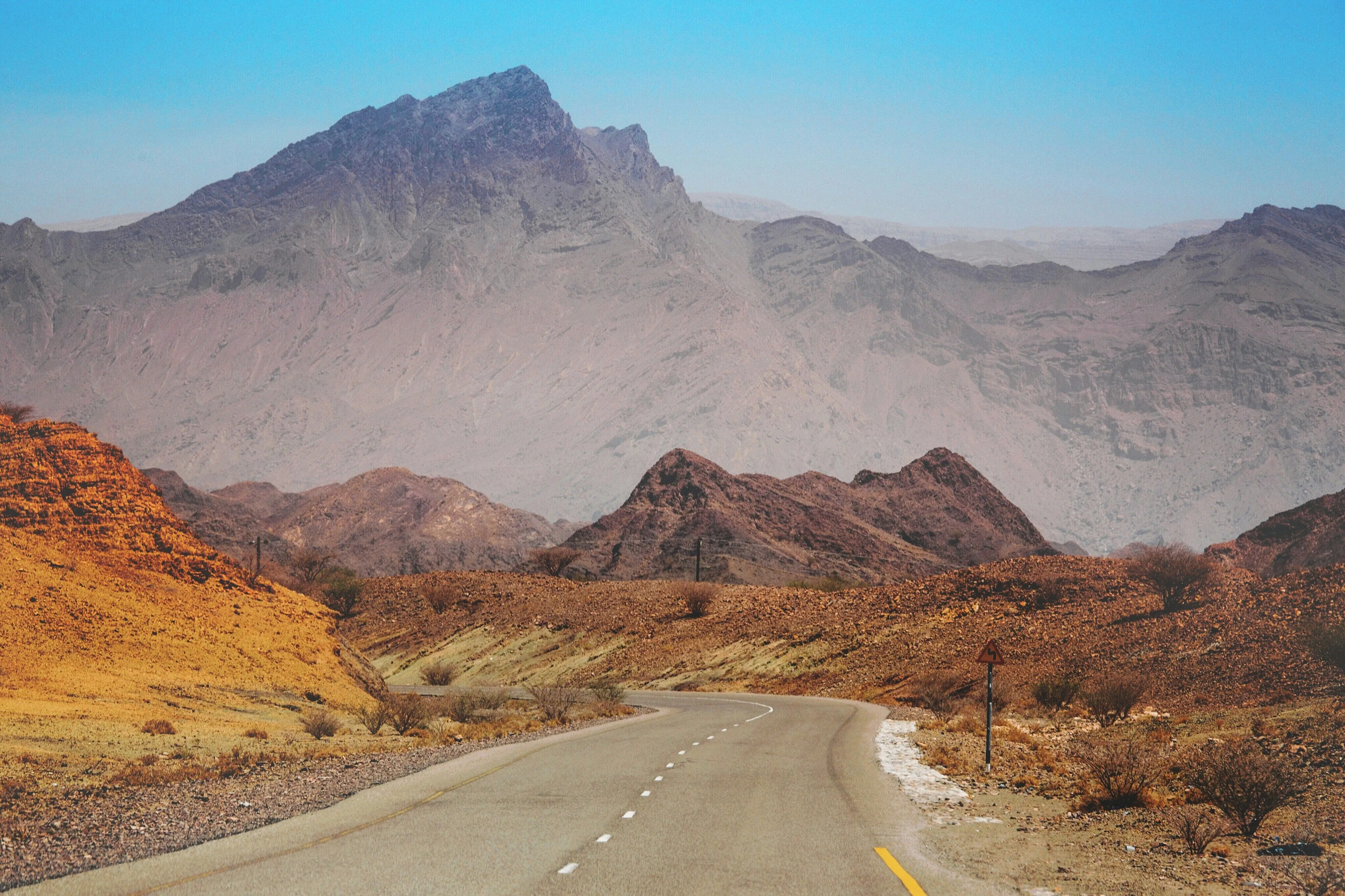 Curved road winding through rocky desert hills with large mountains in the background under a clear blue sky.