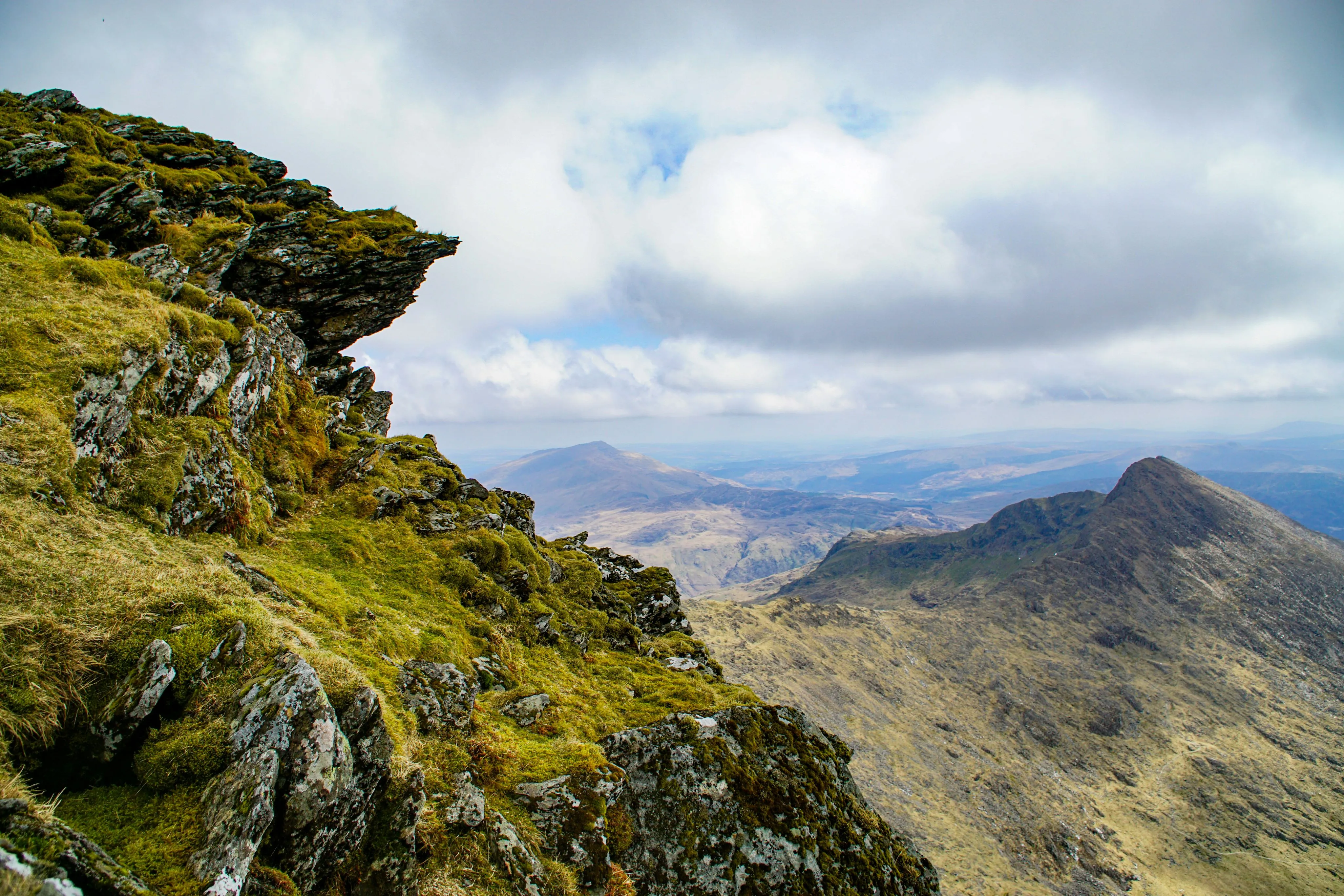 Rocky cliff covered with green moss overlooking distant mountain peaks under a cloudy sky.