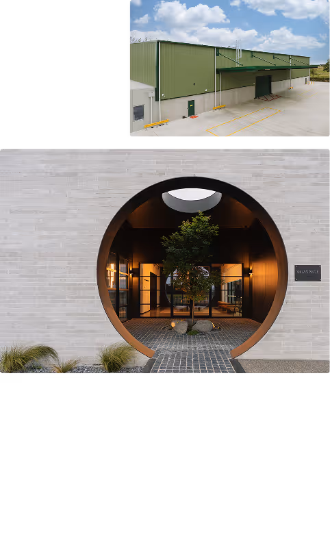 Modern building exterior with a circular entrance showcasing a tree inside under warm lighting, and a green industrial warehouse in the background under a cloudy sky.