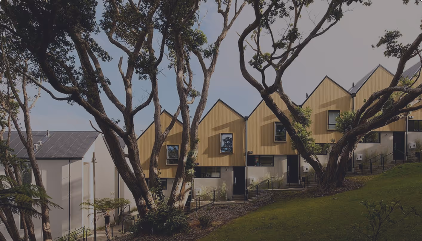 Row of modern townhouses with yellow wooden siding and large windows, partially obscured by tall trees in the foreground.