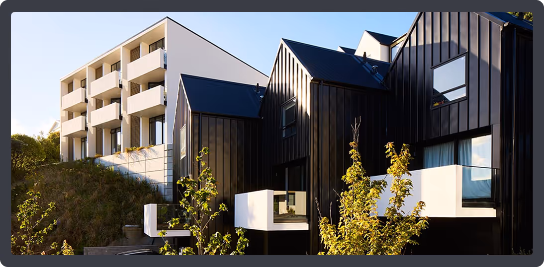 Modern residential buildings with a white multi-story block and adjacent black houses featuring white balconies, surrounded by greenery.