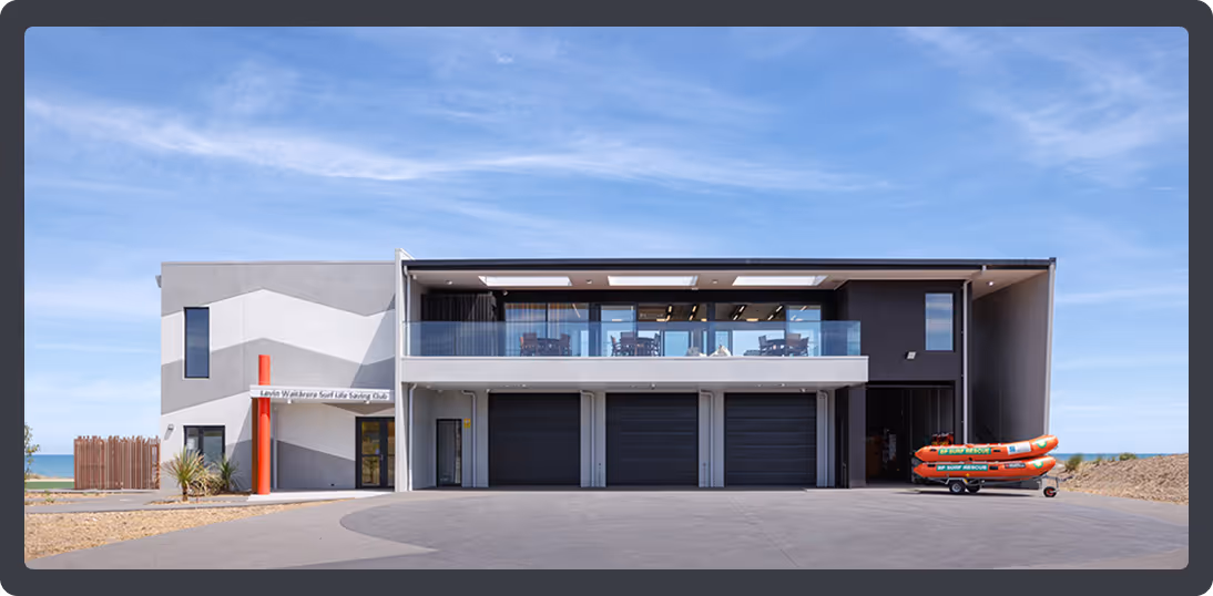 Modern two-story surf lifesaving club building with glass balcony, three black garage doors, and rescue boats on a trailer outside.
