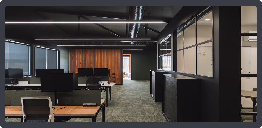 Modern office interior with rows of desks, computer monitors, black chairs, and large windows with blinds.