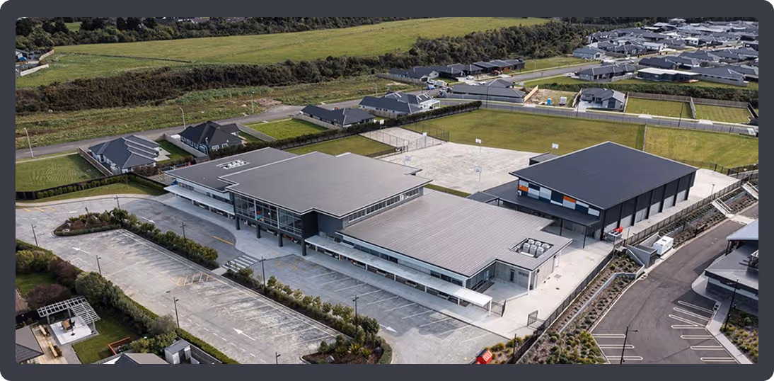 Aerial view of a modern school campus with multiple buildings, parking lots, sports courts, and surrounded by residential houses and green fields.