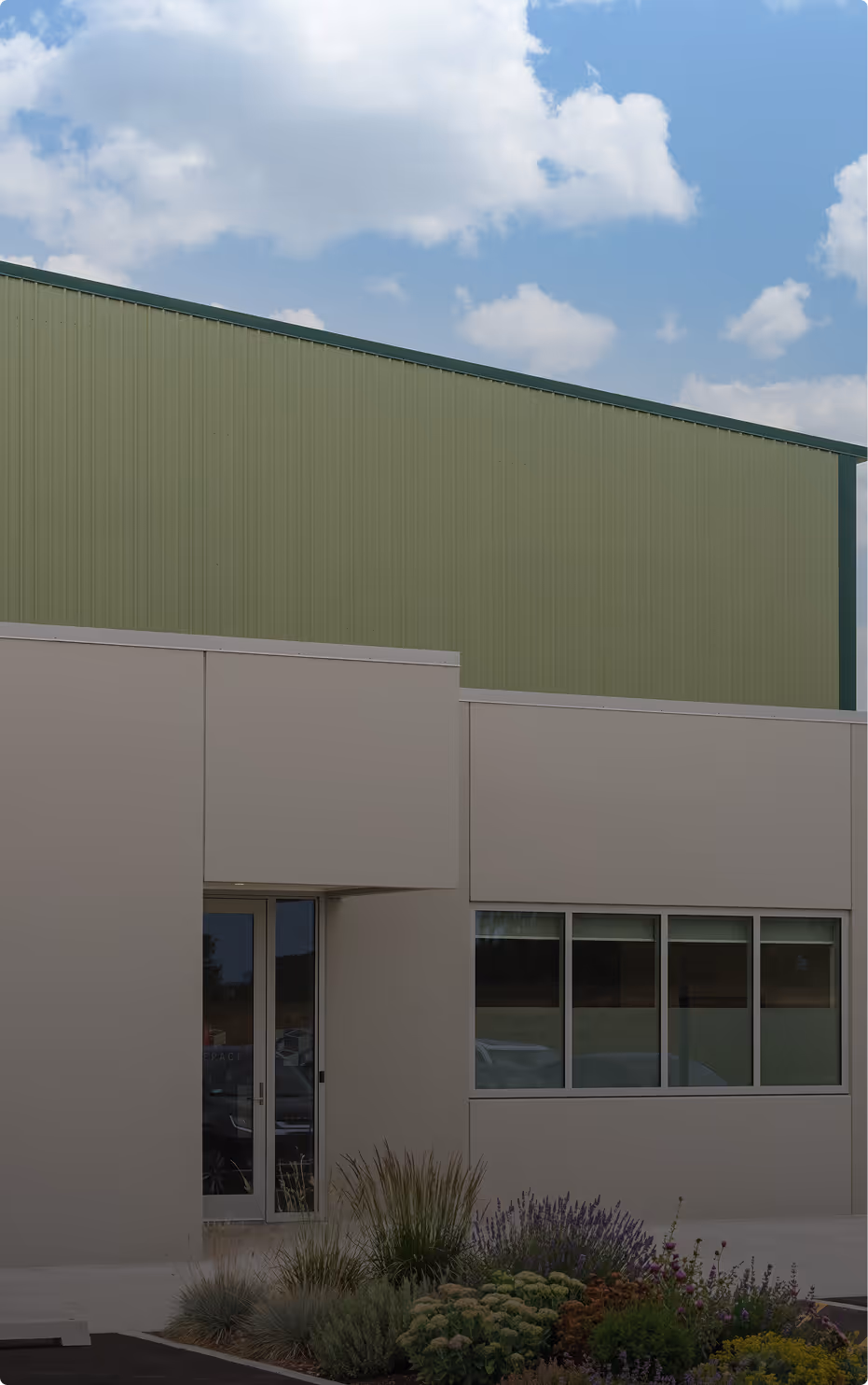 Modern industrial building with beige entrance and large green corrugated metal upper wall under a partly cloudy sky.