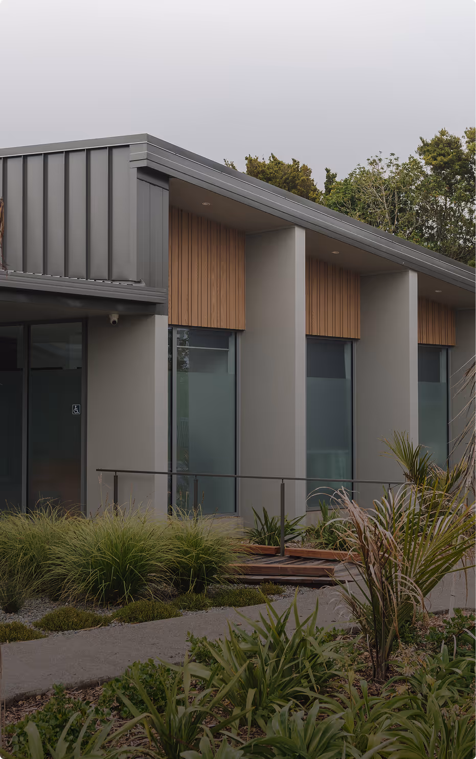 Modern building exterior with large vertical windows, gray walls, wooden panels, and landscaped greenery under an overcast sky.