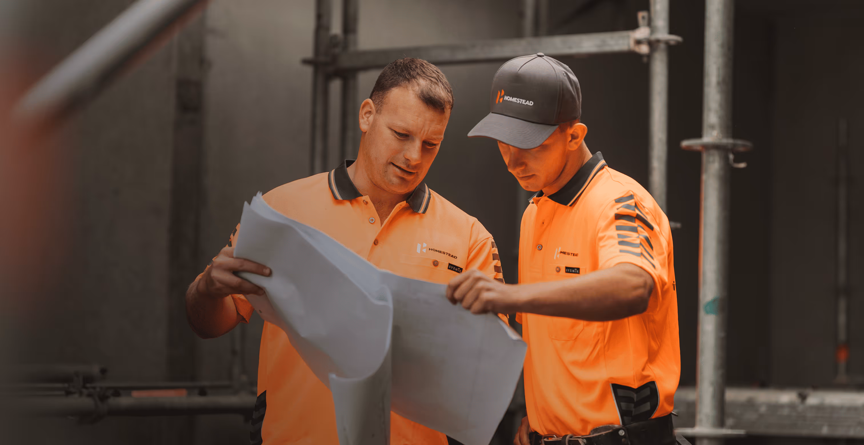 Two construction workers in orange shirts and one wearing a cap reviewing blueprints at a worksite.