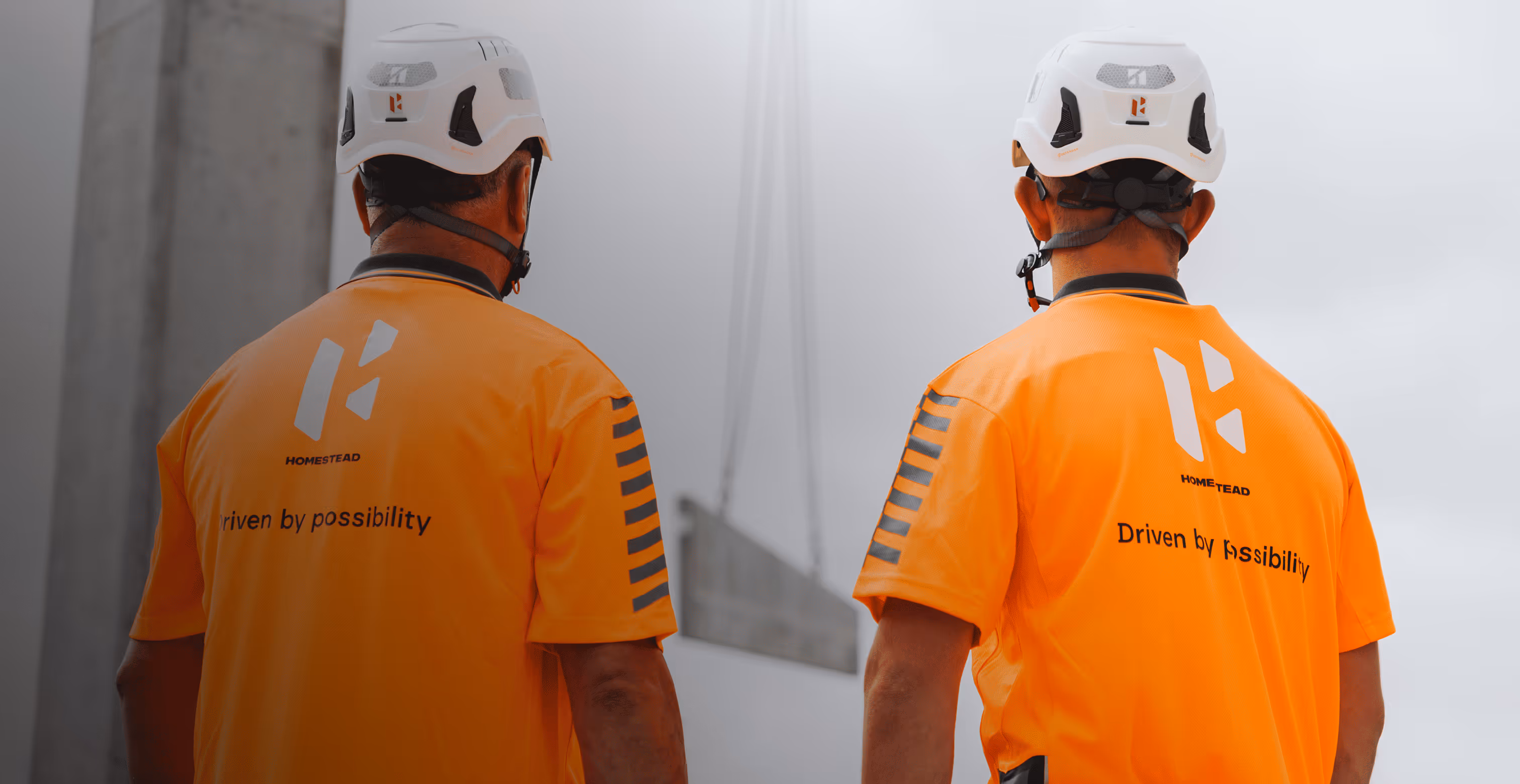 Two workers wearing white helmets and bright orange shirts with the text 'Driven by possibility' standing with their backs facing the camera.