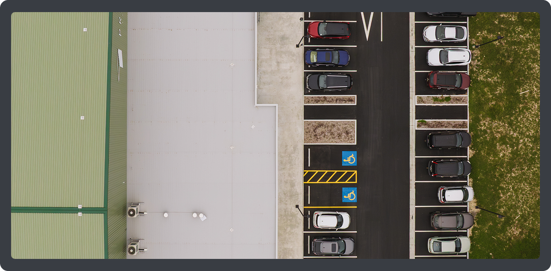 Aerial view of a parking lot with a building on the left, several parked cars, and two handicap parking spaces marked with blue and yellow lines.