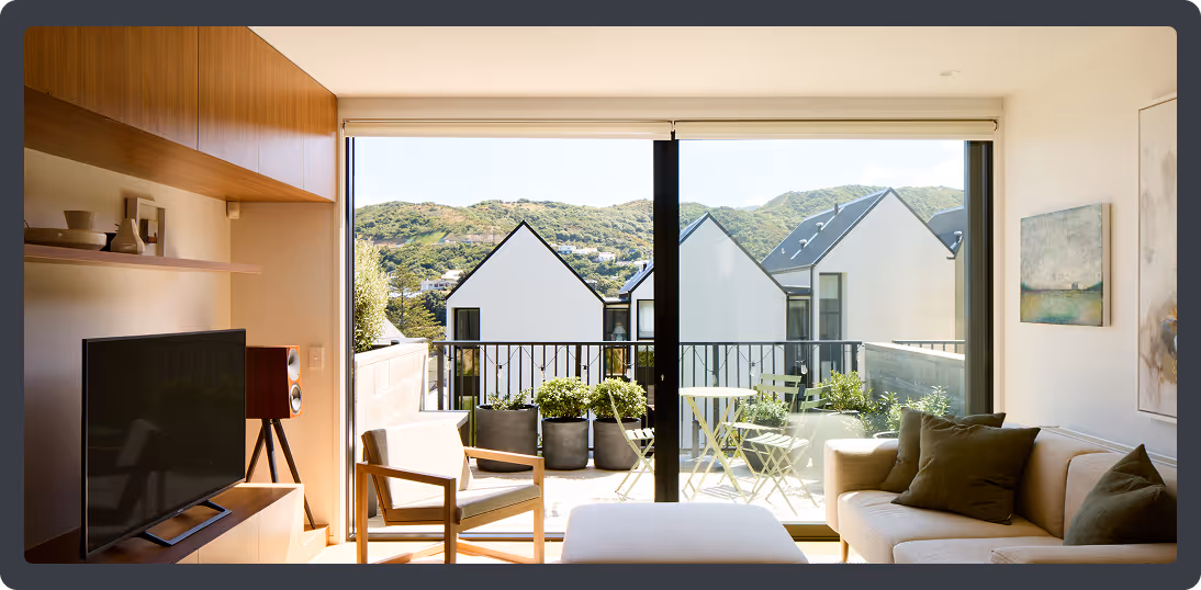 Modern living room with beige sofa, chair, TV, and large glass doors opening to a balcony with plants and a view of white houses and green hills.