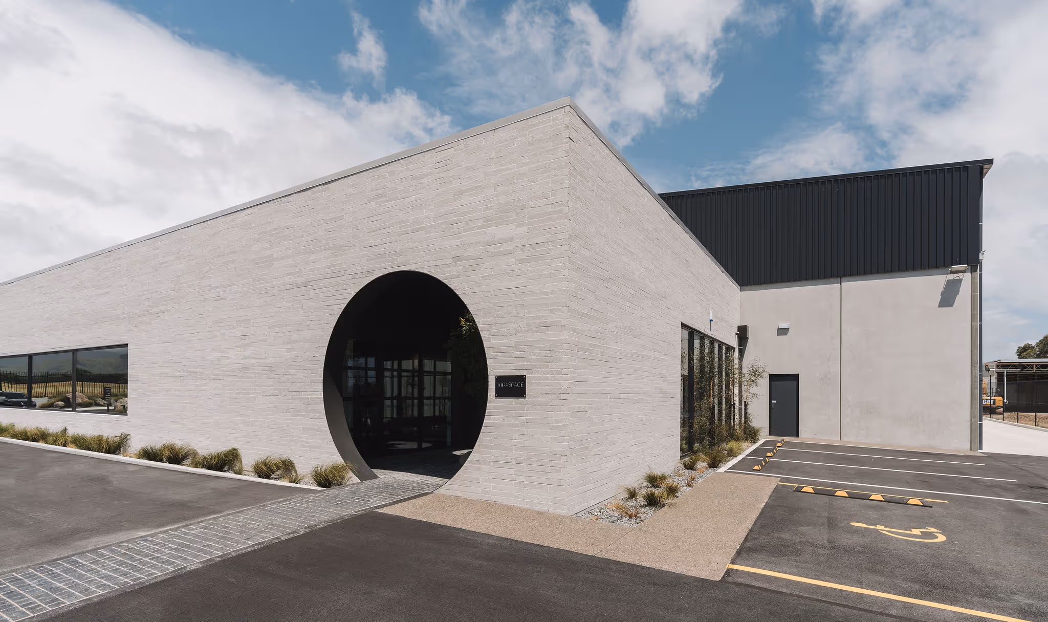 Modern building exterior with light brick walls, large circular entrance, and adjacent parking spaces including a disabled parking spot.