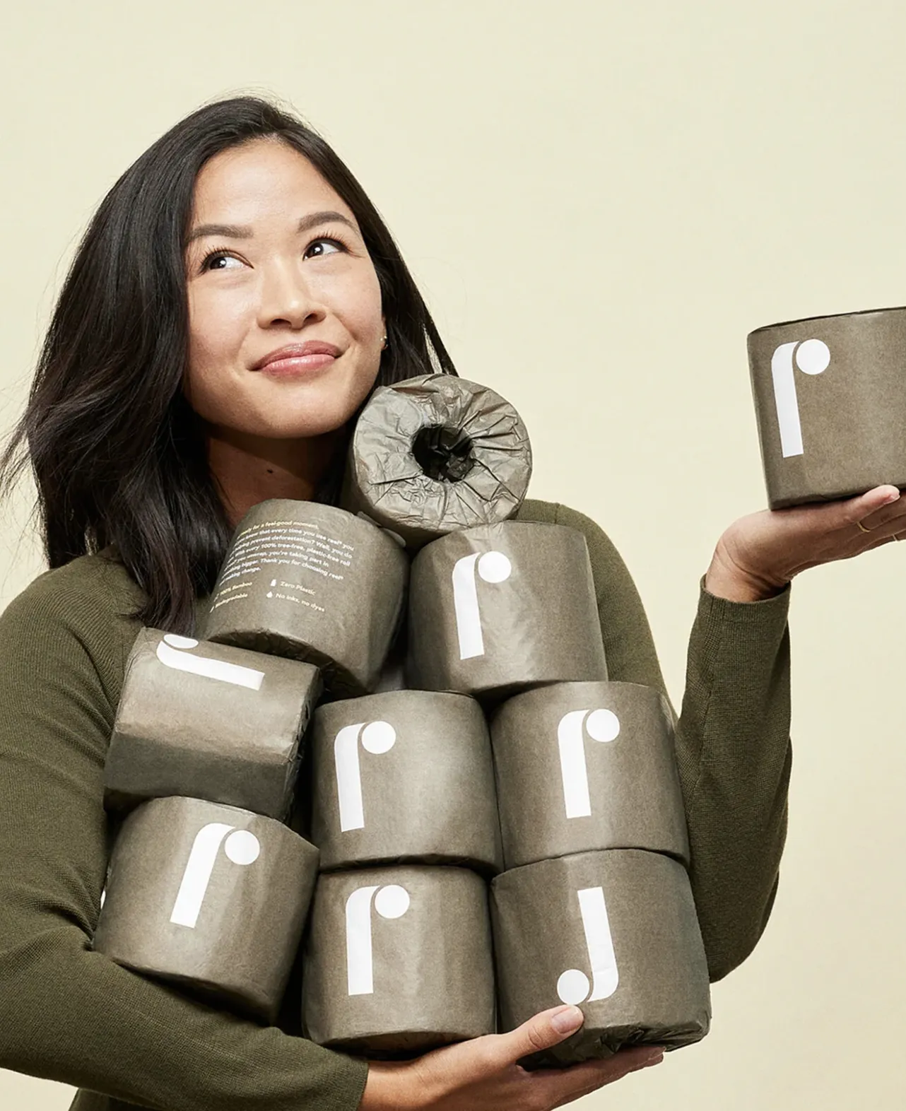 Woman holding a stack of brown recycled toilet paper rolls with a white lowercase 'r' logo on a beige background.