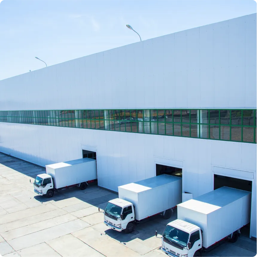 Three white delivery trucks parked at loading dock doors of a large warehouse with green-tinted windows.