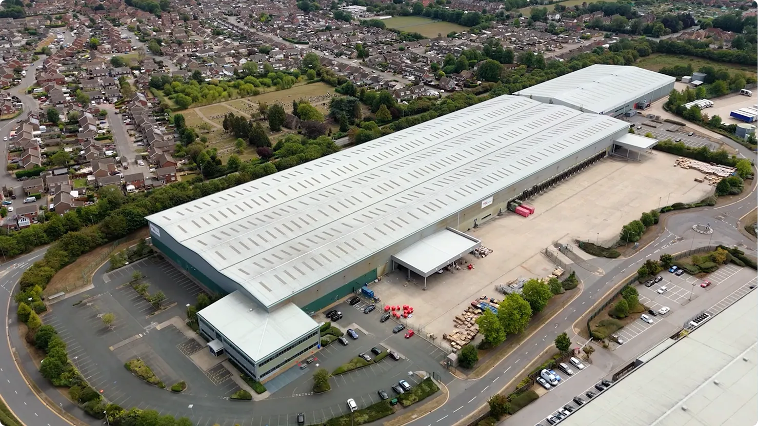 Aerial view of a large industrial warehouse with parking lots, surrounded by residential houses and greenery.