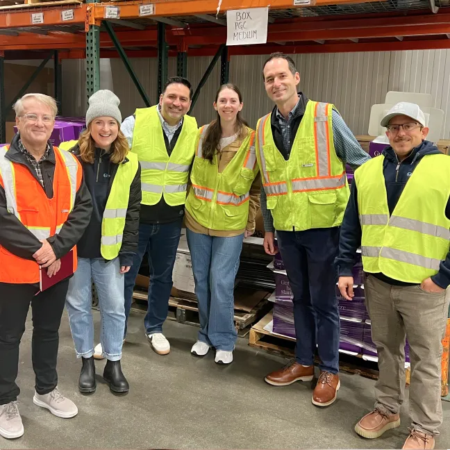 Group of six people wearing safety vests standing inside a warehouse with pallets and boxes.