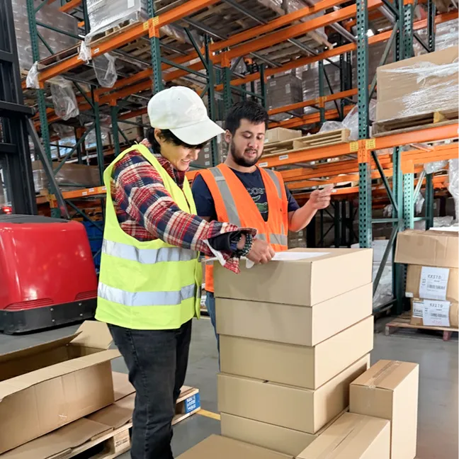 Two warehouse workers in safety vests organizing stacked cardboard boxes in an industrial storage area with shelves and pallets.