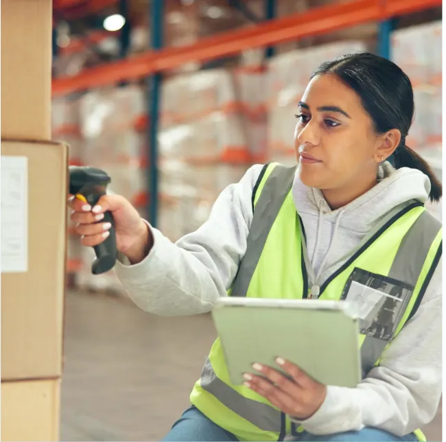 Warehouse worker in a high-visibility vest scanning boxes with a barcode scanner while holding a tablet.