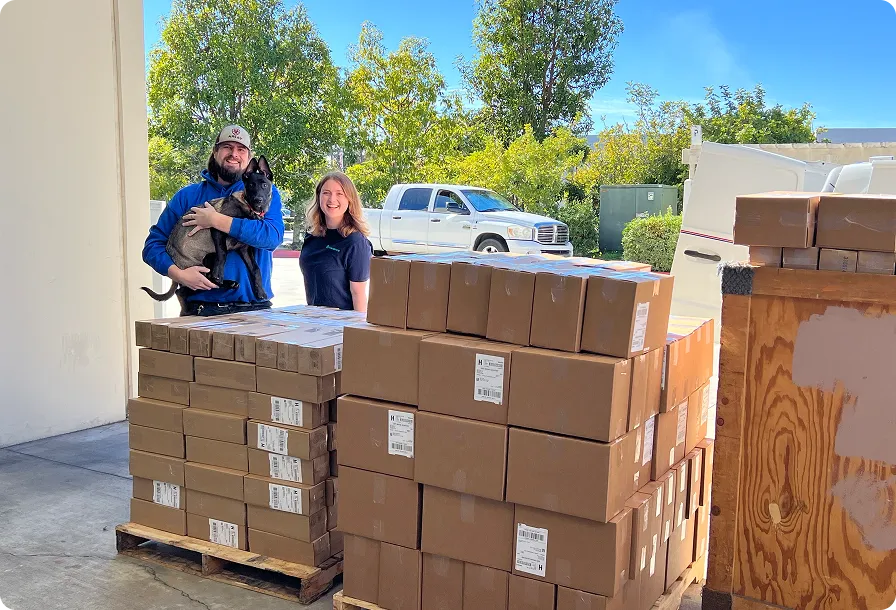 A man holding a dog and a woman standing next to multiple stacked cardboard boxes on pallets outside near a white truck.