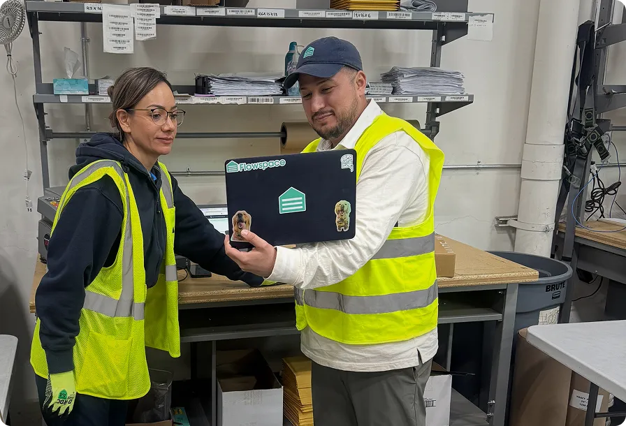 Two warehouse workers wearing high-visibility vests are reviewing information on a laptop with Flowspace stickers in an industrial storage area.
