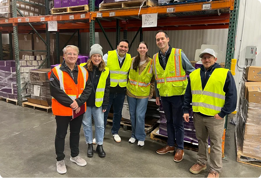 Six people wearing orange and yellow safety vests standing in a warehouse with shelves stocked with boxes and packages.