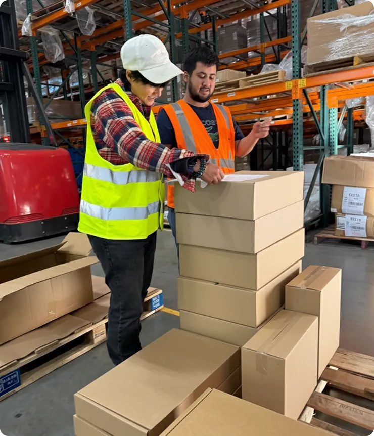 Two warehouse workers in safety vests checking and organizing stacked cardboard boxes on pallets.