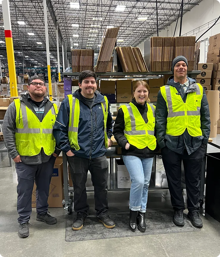 Four warehouse workers wearing yellow safety vests standing and smiling inside a large storage area with stacked cardboard boxes.