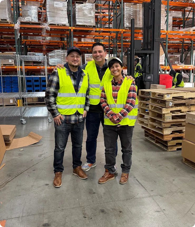 Three warehouse workers wearing yellow safety vests posing and smiling in an industrial storage area with shelves and pallets.